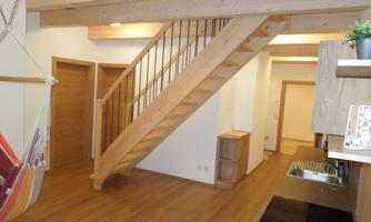 A modern living room with a wooden staircase and open kitchen. The room has light walls and a warm wooden floor.