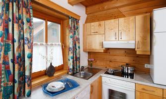 A cozy country-style kitchen with wooden furniture and colorful curtains. The window offers a view outside, while cooking utensils are placed on the countertop.