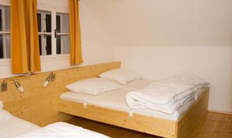 A simple, bright bedroom with two beds. The walls are white, and the windows have orange curtains.