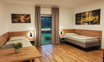 A simple room with two beds and a table. Large windows provide plenty of daylight and a view of the outside.