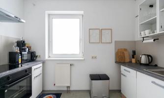 A modern kitchen with bright walls and clean lines. It features a window, cooking appliances, and a waste container.