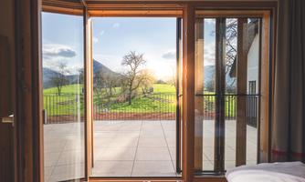 A beautiful view through open French doors onto a green meadow with trees and gentle hills. The sun is shining and creating an inviting atmosphere.