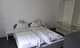 A simple bedroom with two single beds and fresh bedding. In the background, there is an open wardrobe and a nightstand.