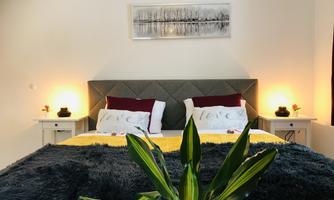 A cozy bedroom with a large bed, soft pillows, and a yellow blanket. Next to the bed are nightstands with lamps and a decorative plant arrangement.