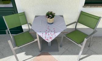 Two green chairs are positioned around a small table with a hexagonal shape. On the table, there is a table mat and a flower in a pot.