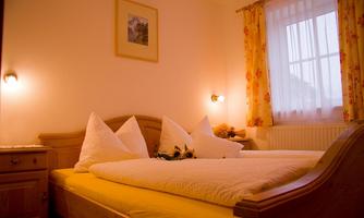 A cozy bedroom with a large double bed and bright pillows. The walls are painted light and decorated with colorful curtains.