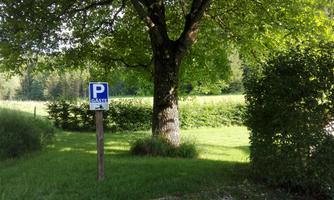 A parking sign for guests is located under a large tree. In the background, green meadows and bushes can be seen.