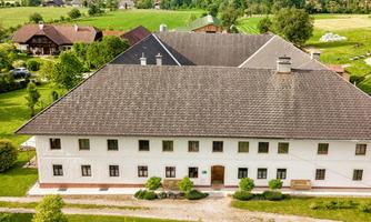 A large white building with a steep roof is situated in a green landscape. Surrounded by trees and meadows, it conveys a rural charm.