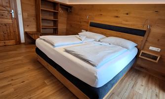 A modern bedroom with a large double bed and wood-paneled walls. The floor is made of wood, and there is a shelf and a bedside lamp.