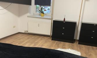 A modern bedroom with wooden floors and minimalist design. There is a television, a wardrobe, and windows with blue blinds.