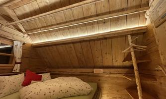 A cozy room in a wooden style with a low, sloping roof. There is a bed with pillows and a wooden shelf.