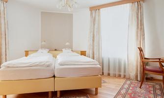 A bright bedroom with two single beds and wooden flooring. Curtains and a chair give the room a cozy atmosphere.