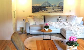 A cozy living room with a gray sofa, a small table, and a large wall painting of mountains. The room is bright and inviting, with a friendly, modern decor.