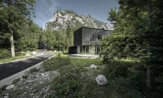 A modern house in a green environment, surrounded by trees and mountains. The road gently passes by the building.