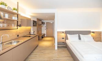 A modern and bright room with a kitchenette and a cozy bed. The decor is simple and inviting, with wooden shelves and warm lighting.