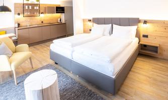 A modern bedroom with a comfortable double bed and wooden elements. The decor is bright and inviting, featuring a kitchenette and a cozy seating area.