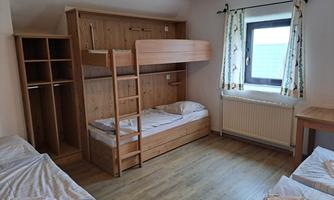 A simple room with a bunk bed and two single beds. There are wooden shelves and a window with curtains.