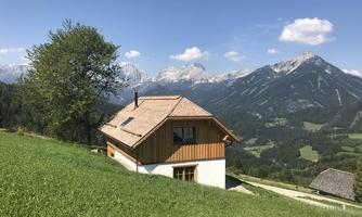 A pretty house sits on a meadow with a view of the mountains. The sky is blue and there are some clouds.