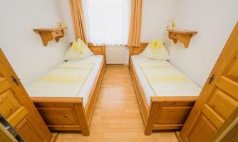 A simple room with two single beds and light wooden shelves. The walls are painted in neutral colors and a window lets in plenty of daylight.