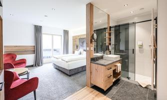 A modern hotel room with a comfortable bed and an adjacent bathroom. Red armchairs and large windows create an inviting atmosphere.