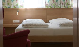 A simple bedroom with a double bed and fresh bed linens. Bright flowering curtains hang on the wall, and there is a red armchair.
