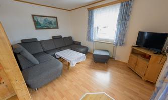 A cozy living room with a gray sofa and a coffee table. A television and a sideboard are also visible, as well as a window with curtains.