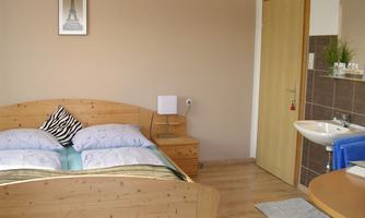 A cozy bedroom with a wooden bed and blue pillows. In the background, there is a bathroom area with a sink and a towel.