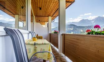 A beautiful balcony with a view of the mountains. On the table, there is a tablecloth and some flowers are blooming in the window boxes.