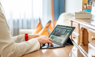 A person is working on a laptop at a table. In the background, pillows and decorative items are visible.