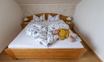 A cozy bedroom with a large wooden bed and colorful pillows. A small child is lying relaxed on the bedspread.