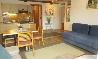 A cozy living room with a blue sofa and a dining table. The modernly furnished kitchen is visible in the background.