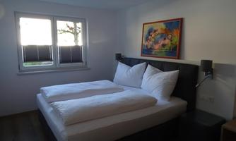 A modern bedroom with a large double bed and white pillows. A colorful picture hangs on the wall, and daylight comes through the window.
