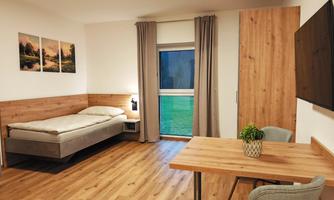 A bright room with wooden floors, a single bed, and a table. Pictures hang on the walls, and the window lets in a lot of light.