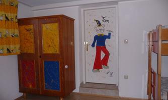 A child-friendly room with colorful curtains and a carved wardrobe. The door is decorated with a cheerful clown motif.