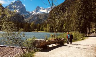 A picturesque mountain landscape with a clear lake and snow-capped peaks. Two people are walking along a path, surrounded by trees and natural beauty.