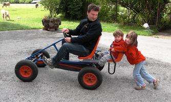 A man is driving a go-kart with two children sitting on it. The children are laughing and wearing red sweaters.
