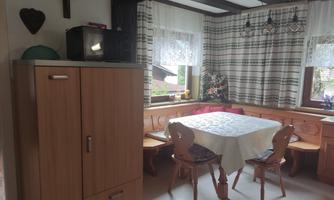 A cozy kitchen with a wooden table and two chairs. In the background, there is a refrigerator and a microwave, surrounded by curtains and window light.