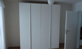 A large white wardrobe stands against the wall in a bright room. Next to it is an open door and a window with curtains.