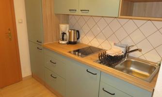 A modern kitchen with green cabinets and a wood countertop. The amenities include a kettle, a sink, and a stove.