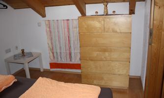 A cozy bedroom with a wooden dresser and a small table. The walls are painted in light colors and a decorative cloth hangs on the wall.