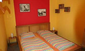 A cozy bedroom with a double bed and colorful blankets. The walls are yellow and red, with decorative pictures and shelves.