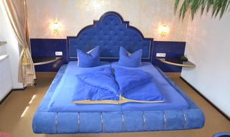 A modern bedroom with a large, blue bed and stylish wall design. The room is brightly lit and has a cozy atmosphere.