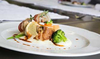 An artistically arranged plate with fried meat wrapped in bacon, accompanied by a side of broccoli and melon. A creamy sauce completes the dish.