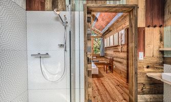 The image shows a modern bathroom with a shower and wood paneling. Through the open door, a cozy living space with wooden flooring and a chair is visible.