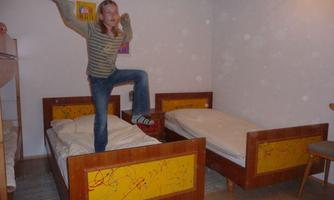 A girl is standing on a bed in a simple room. There are two beds with colorful patterns and a chair.