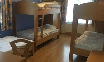 A simple room with bunk beds and a single bed. There is a table and chairs as well as large windows that let in light.
