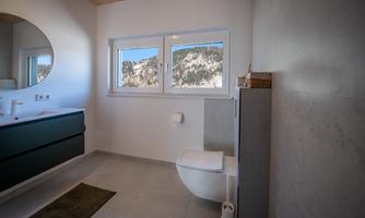 A modern bathroom with a large mirror and a window that offers a view of the mountains. The walls are bright and simple, the decor is minimalist.
