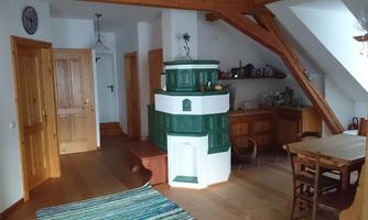 A cozy room with wooden floors and a green stove in the center. There are wooden furniture and plenty of daylight.