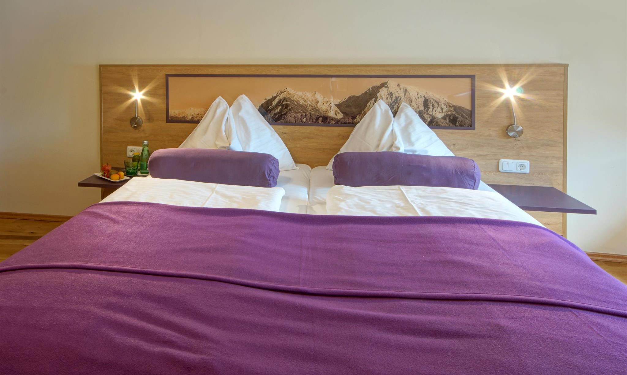 A cozy bedroom with a large bed and purple pillows. On the wall, there is a decorative picture of mountains.