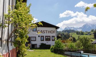 An inn with a beautiful facade and a green surrounding. In the background, the mountains and a blue sky are visible.